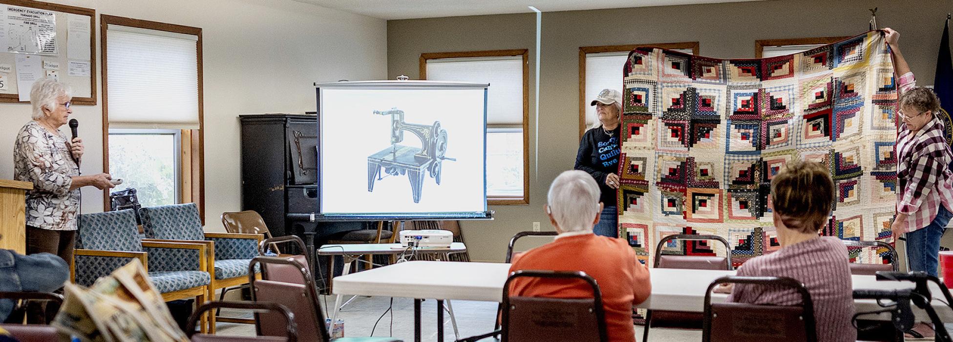 Yvonne Hollenbeck, left, explains quilting from the Civil War to today’s quilts which she has made. Brenda Hofeldt of Millboro, and Kay Evans of Storm Lake, IA, are the quilt holders of these precious stories of the past and present. On the screen is a hand crank sewing machine, first available in the 1860s. The Log Cabin quilt pictured was made by Yvonne’s great-great grandmother. Photo by Laura Vroman