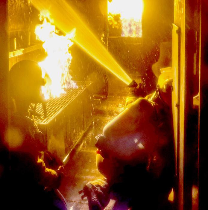cation, and executing a plan to knock the flames down. No pressure?!? At left, inside the burning trailer, three firefighters man the hose, working together to douse